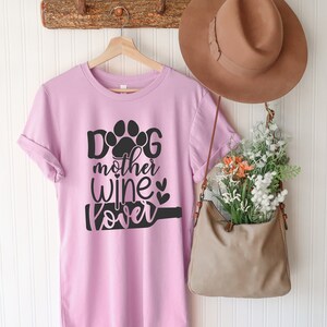 May include: A pink t-shirt with a black paw print and the text "Dog mother wine lover" printed on it. The shirt is hanging on a wooden rack with a brown hat and a brown purse.