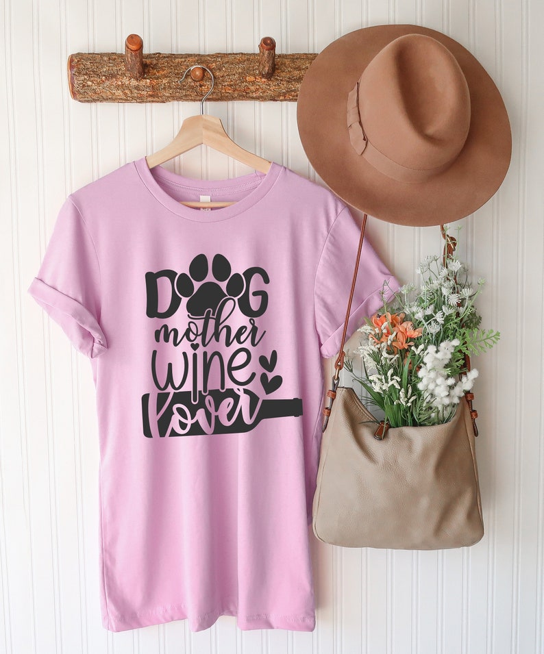May include: A pink t-shirt with a black paw print and the text "Dog mother wine lover" printed on it. The shirt is hanging on a wooden rack with a brown hat and a brown purse.