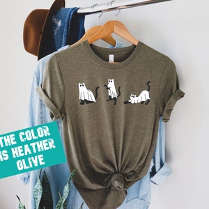 May include: A heather olive t-shirt with a graphic of three cats dressed as ghosts. The text "THE COLOR IS HEATHER OLIVE" is on a teal rectangle.