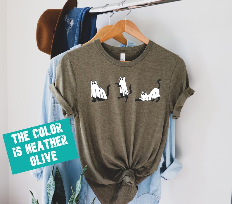 May include: A heather olive t-shirt with a graphic of three cats dressed as ghosts. The text "THE COLOR IS HEATHER OLIVE" is on a teal rectangle.