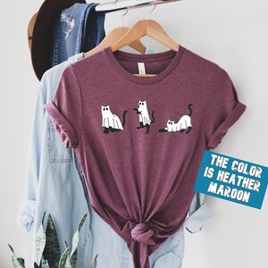 May include: A heather maroon t-shirt with a graphic of three cats dressed as ghosts. The text "THE COLOR IS HEATHER MAROON" is on a blue rectangle.