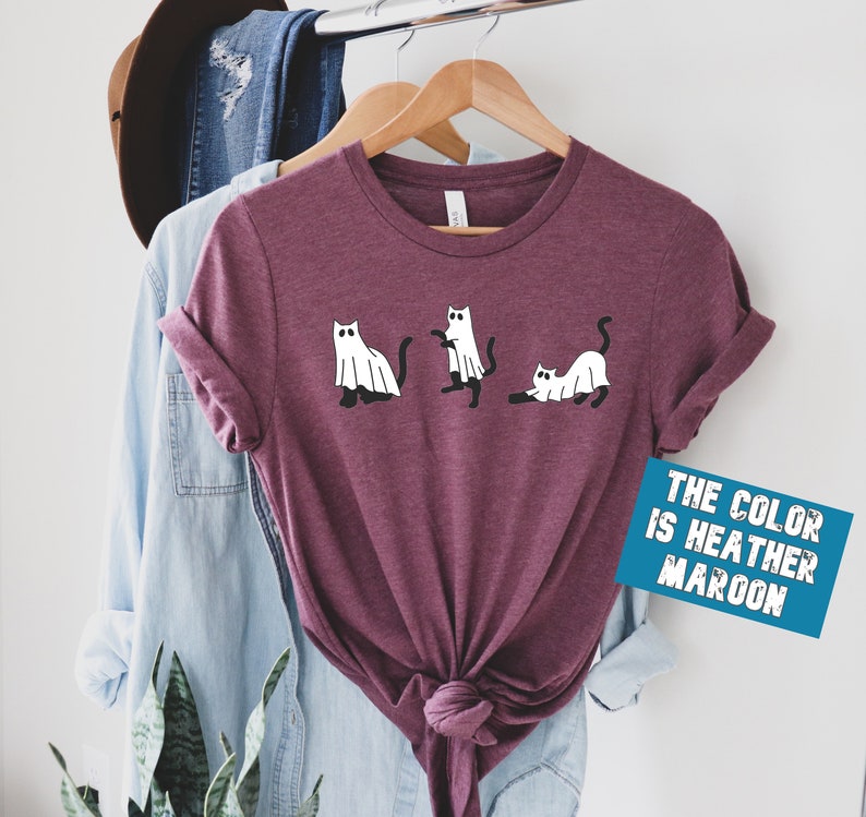 May include: A heather maroon t-shirt with a graphic of three cats dressed as ghosts. The text "THE COLOR IS HEATHER MAROON" is on a blue rectangle.