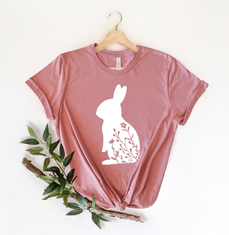 May include: A dusky pink t-shirt with a white rabbit design and floral details. The tee is displayed on a wooden hanger against a white backdrop, with a sprig of foliage in the lower left corner. The t-shirt is knotted at the bottom.