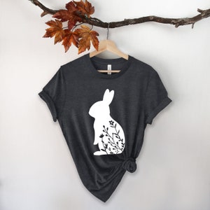 May include: Dark grey t-shirt with a white rabbit silhouette and floral design. The tee is displayed on a wooden hanger. A branch with autumnal leaves hangs above. The background is white.