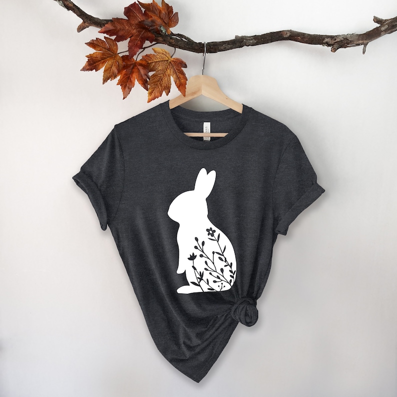May include: Dark grey t-shirt with a white rabbit silhouette and floral design. The tee is displayed on a wooden hanger. A branch with autumnal leaves hangs above. The background is white.