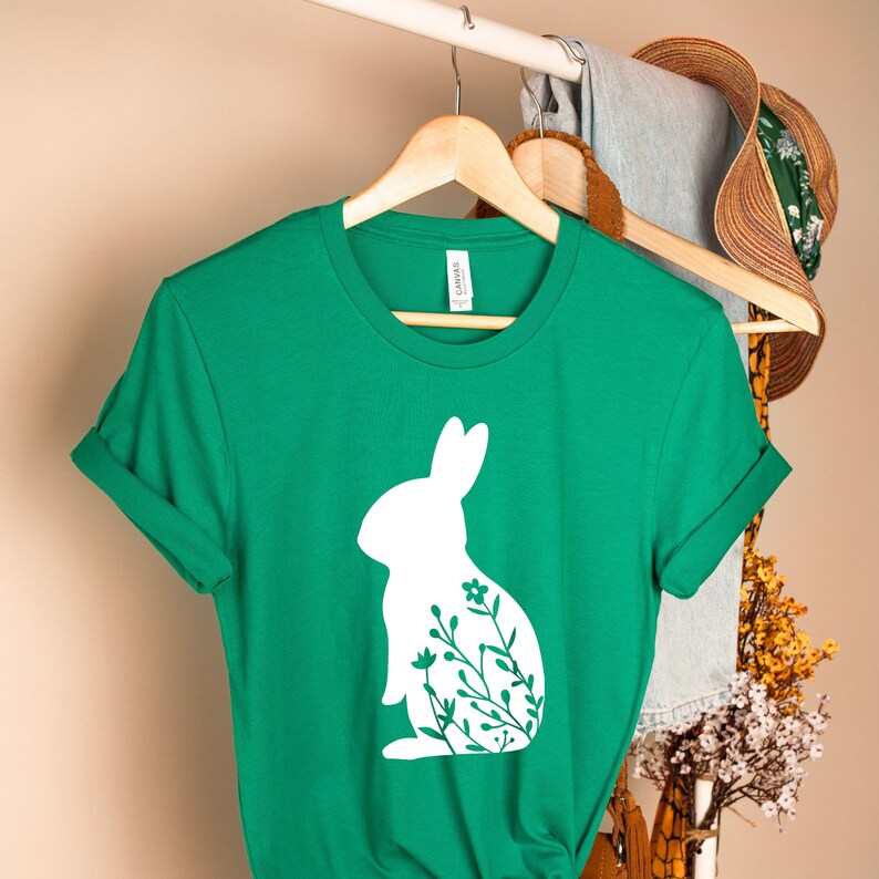 May include: A green t-shirt featuring a white rabbit silhouette filled with floral details. The tee is displayed on a wooden hanger. Other garments and a straw hat are visible in the background.