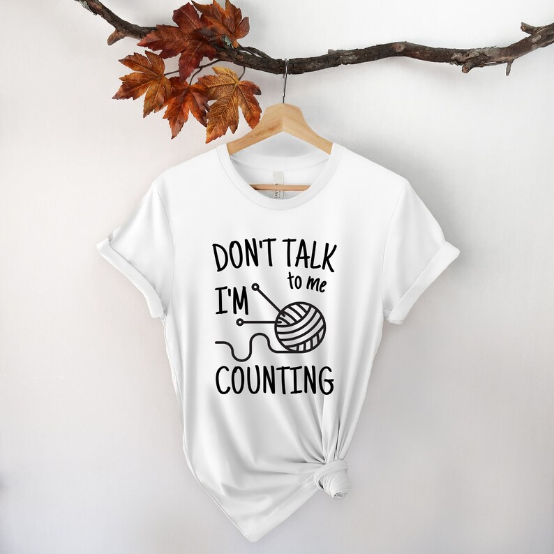 May include: White t-shirt with black text that reads "Don't talk to me I'm counting" with a black and white illustration of a ball of yarn and knitting needles.