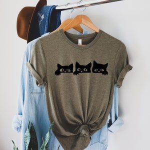 May include: A green t-shirt with a graphic of three black cats peeking over the top. The cats have white eyes and are looking to the right.