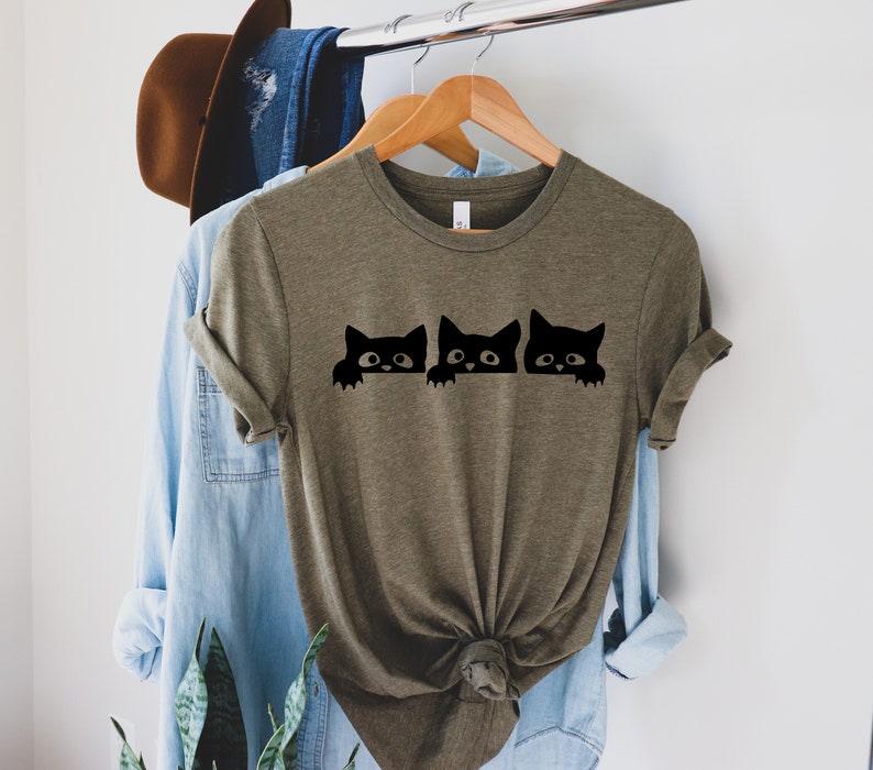 May include: A green t-shirt with a graphic of three black cats peeking over the top. The cats have white eyes and are looking to the right.