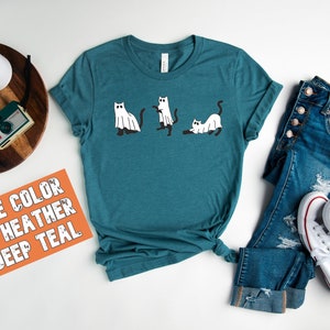 May include: A teal blue t-shirt with a graphic of three cats dressed as ghosts. The text "THE COLOR IS HEATHER DEEP TEAL" is on a bright orange card.