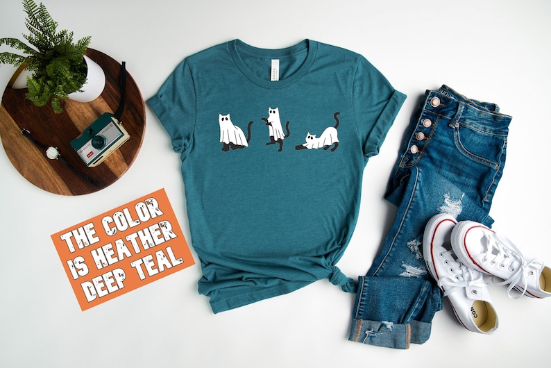 May include: A teal blue t-shirt with a graphic of three cats dressed as ghosts. The text "THE COLOR IS HEATHER DEEP TEAL" is on a bright orange card.