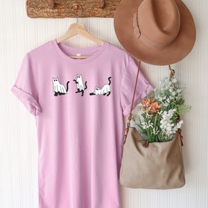 May include: A light purple t-shirt with a graphic of three white cats dressed as ghosts. The shirt is hanging on a wooden hanger with a brown hat and a brown bag hanging next to it.