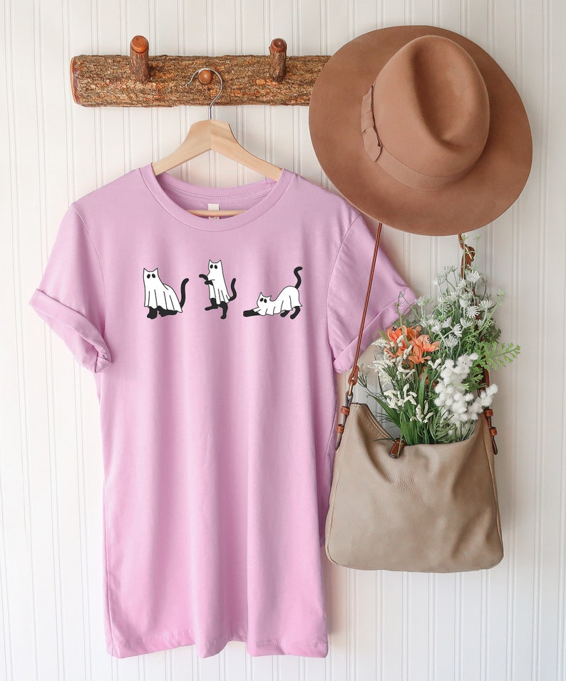 May include: A light purple t-shirt with a graphic of three white cats dressed as ghosts. The shirt is hanging on a wooden hanger with a brown hat and a brown bag hanging next to it.