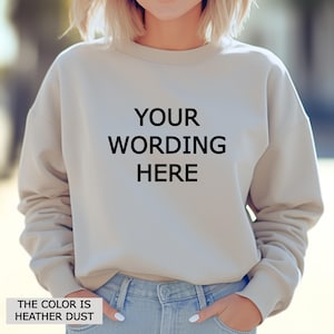 Peut inclure: Un sweat-shirt beige avec le texte "YOUR WORDING HERE" en noir. Le sweat-shirt est de couleur neutre et a des manches longues. Le texte "THE COLOR IS HEATHER DUST" est en bas de l'image.