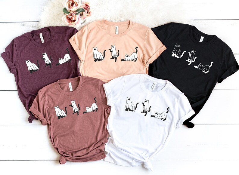 May include: Five t-shirts with a graphic of cats dressed as ghosts. The shirts are in various colors, including maroon, peach, black, brown, and white.