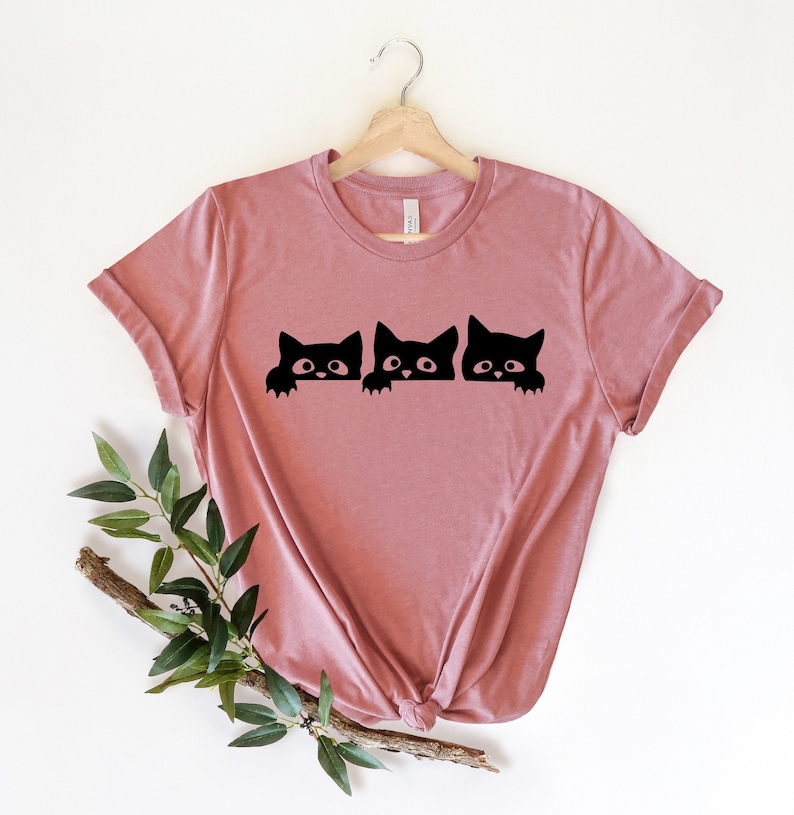 May include: A dusty rose t-shirt with a graphic of three black cats peeking over a surface. The cats have white eyes and are looking at the viewer.
