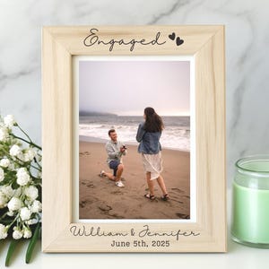 May include: A light-colored wooden picture frame with the word "Engaged" and two hearts at the top. The frame holds a photo of a beach proposal. The names "William & Jennifer" and the date "June 5th, 2025" are printed at the bottom.