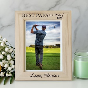 May include: A light-colored wooden picture frame with the text "BEST PAPA BY PAR" and "Love, Olivia" engraved on it. The frame holds a photo of a person playing golf. A yellow rose and a green candle are next to the frame.