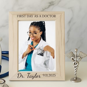 May include: A wooden picture frame with the text "FIRST DAY AS A DOCTOR" and "Dr. Taylor 9.11.2025" contains a photo of a smiling person wearing glasses and a white coat. A blue stethoscope and a silver caduceus statue are also present.