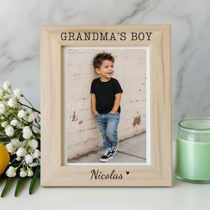May include: A light-colored wooden picture frame featuring the engraved text "GRANDMA'S BOY" at the top and "Nicolas" with a small heart symbol at the bottom. The frame holds a photograph of a young child wearing a black t-shirt and blue jeans.