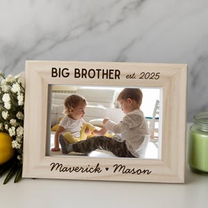 May include: A light-colored wooden picture frame with the words "BIG BROTHER est. 2025" at the top and "Maverick ♥ Mason" at the bottom. The frame holds a photo of two young children. A yellow rose and a green candle are also present.