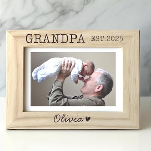 May include: A wooden photo frame with the text "Grandpa Est. 2025" and "Olivia" with a heart symbol. The frame contains a photo of a man holding a baby.