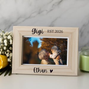 May include: A light-colored wooden picture frame with the text Gigi EST.2026 engraved on the top border and the name Ethan with a heart symbol on the bottom border. The frame holds a photograph of an adult and a baby outdoors in autumn.
