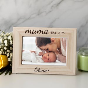 May include: A light-colored wooden picture frame with the word "mama" and "EST. 2025" printed on it. The frame holds a photo of a baby and a person. A yellow rose and a green candle are next to the frame.