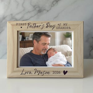May include: A light-colored wooden picture frame with a photo of a man holding a baby. The frame has the text "First Father's Day as my Grandpa" and "Love, Mason 2026" with a heart symbol.