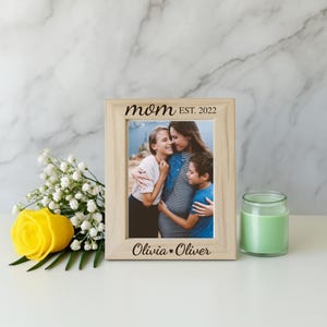 May include: A light wooden picture frame with the words "mom EST. 2022" and "Olivia &bull; Oliver" engraved on it. The frame holds a photo of a mother and two children. A yellow rose and a green candle are also in the image.