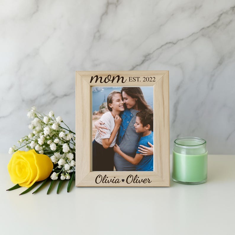 May include: A light wooden picture frame with the words "mom EST. 2022" and "Olivia &bull; Oliver" engraved on it. The frame holds a photo of a mother and two children. A yellow rose and a green candle are also in the image.
