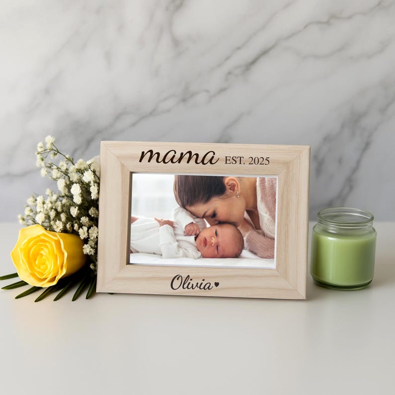 May include: A light-colored wooden picture frame with the word "mama" and "EST. 2025" printed on it. The frame holds a photo of a baby and a person. A yellow rose and a green candle are next to the frame.
