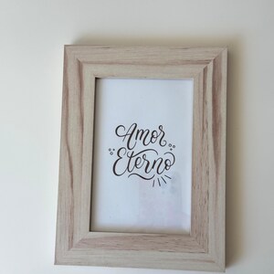 May include: A rectangular wooden picture frame with a light brown finish. The frame holds a white print with the words "Amor Eterno" in elegant black script. The frame is set against a plain white background.