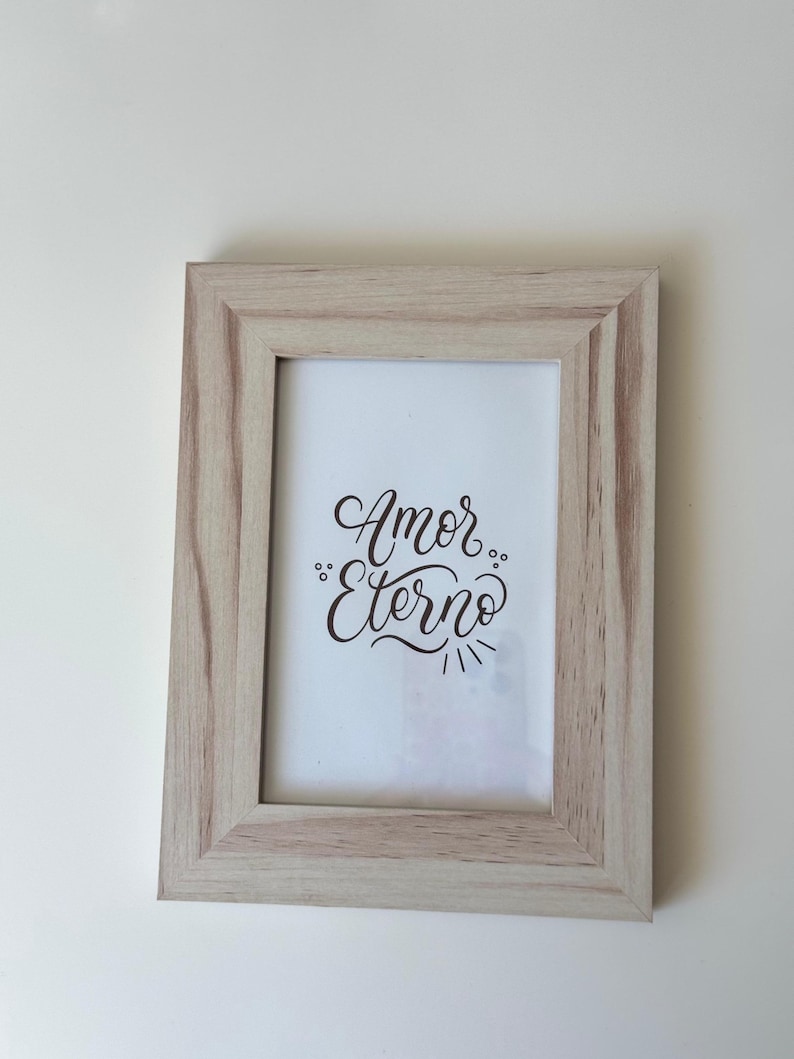 May include: A rectangular wooden picture frame with a light brown finish. The frame holds a white print with the words "Amor Eterno" in elegant black script. The frame is set against a plain white background.