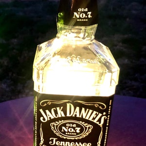 May include: A clear glass Jack Daniel's whiskey bottle with a black label. The label features the words "Jack Daniel's" and "Old No. 7 Brand". The bottle is filled with a light-coloured liquid and has a cork stopper.