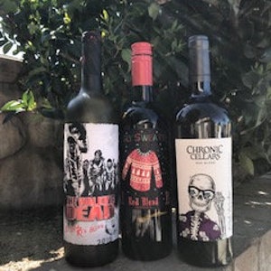 Puede incluir: Tres botellas de vino con etiquetas únicas se alzan contra un fondo de follaje verde. Las etiquetas presentan ilustraciones: un diseño de zombis, un suéter festivo y un esqueleto. Las botellas son oscuras, con detalles en rojo y blanco.