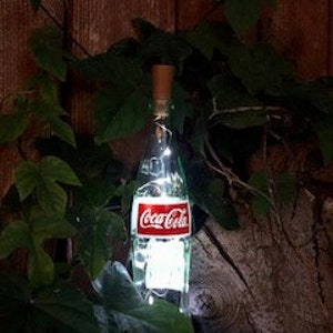 May include: A repurposed Coca-Cola bottle with a cork stopper and string lights inside. The bottle features the classic red and white Coca-Cola label. The bottle is surrounded by green leaves and set against a wooden background.