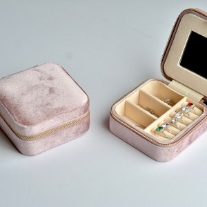 以下が含まれることがあります： ジッパー留めと鏡が付いたピンクのベルベット製のジュエリーボックス。ボックスには、指輪やその他の小さなジュエリーを収納するための複数のコンパートメントがあります。