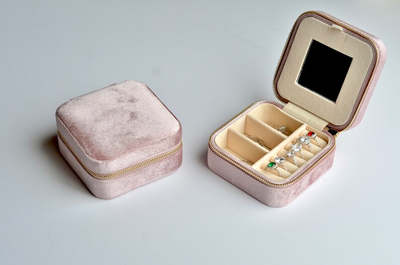 以下が含まれることがあります： ジッパー留めと鏡が付いたピンクのベルベット製のジュエリーボックス。ボックスには、指輪やその他の小さなジュエリーを収納するための複数のコンパートメントがあります。