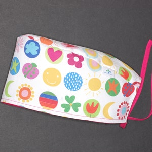 May include: White surgical scrub cap with a colorful pattern of flowers, hearts, ladybugs, and smiley faces. The cap has a pink trim and matching ties for securing. The design is playful and bright.