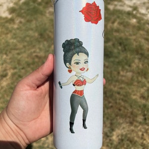 May include: A white tumbler with a glittery finish features a cartoon illustration of a woman with dark hair, wearing a red top and black pants. The tumbler also has a red rose illustration.