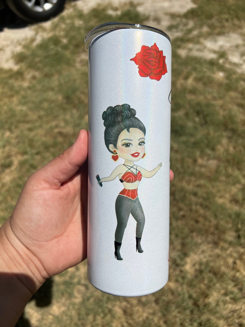 May include: A white tumbler with a glittery finish features a cartoon illustration of a woman with dark hair, wearing a red top and black pants. The tumbler also has a red rose illustration.