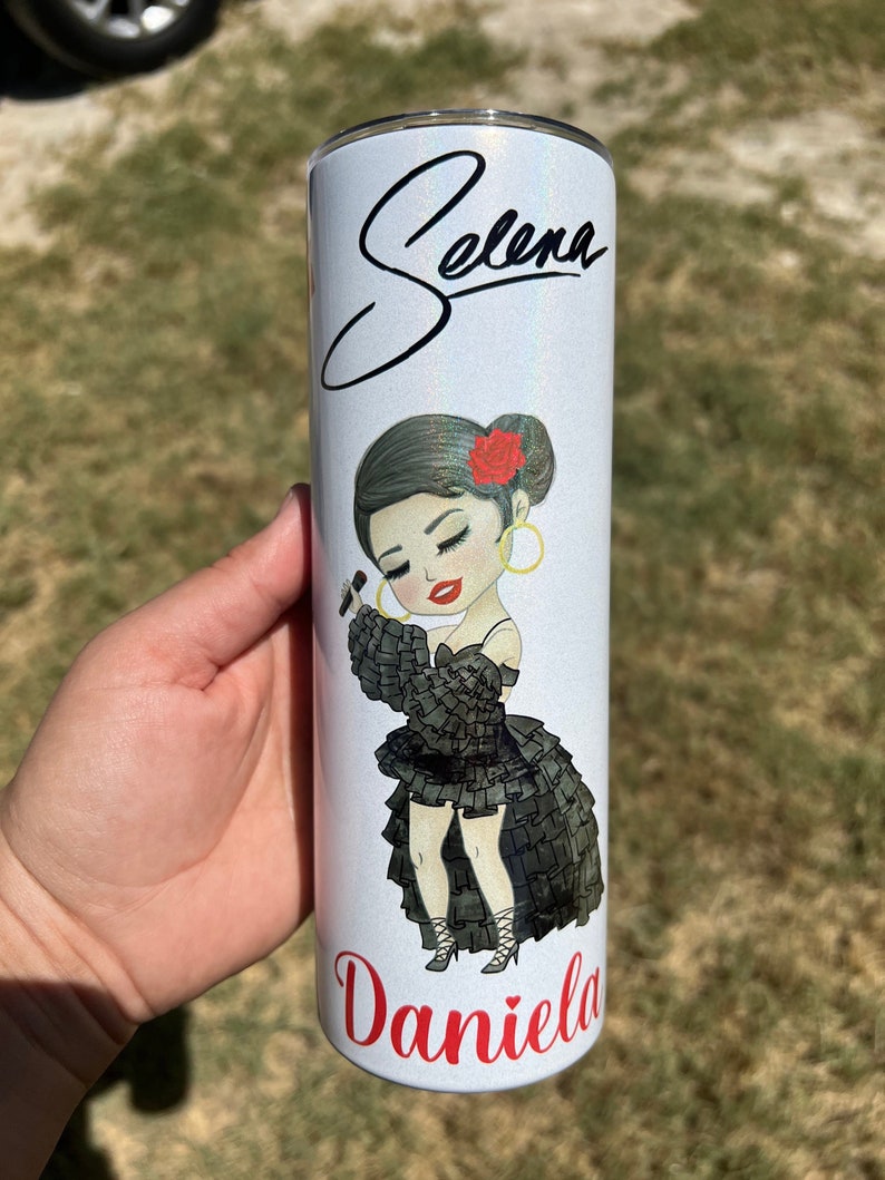 May include: A white stainless steel tumbler with a glittery finish. It features a cartoon illustration of Selena with a microphone and the text "Selena" and "Daniela" in red.