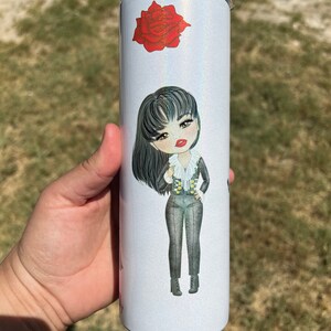 May include: A white stainless steel tumbler with a glittery finish. The tumbler features a cartoon illustration of a woman with long black hair, wearing a black and white outfit. A red rose is also depicted on the tumbler.