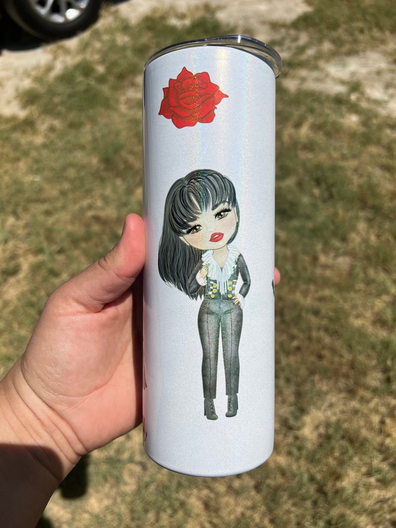 May include: A white stainless steel tumbler with a glittery finish. The tumbler features a cartoon illustration of a woman with long black hair, wearing a black and white outfit. A red rose is also depicted on the tumbler.