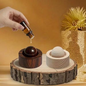Könnte beinhalten: Zwei hölzerne Aromatherapie-Diffusoren, einer dunkelbraun und einer hell, stehen auf einer rustikalen Holzscheibe. Eine Hand gießt Öl aus einer kleinen bernsteinfarbenen Flasche auf den dunkelbraunen Diffusor. Die Diffusoren haben eine runde Oberseite und einen zylindrischen Sockel.