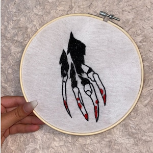 May include: Embroidered hoop art featuring a black and white skeletal hand with red fingernails. The hand is reaching out with claws extended.