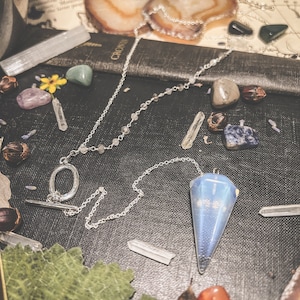 Puede incluir: Una cadena de plata con un péndulo de cristal azul y blanco. La cadena tiene un anillo en la parte superior y está rodeada de varios cristales y piedras.