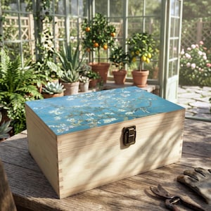 May include: A wooden storage box with a blue lid featuring a floral painting. The box has a brass clasp and is on a wooden table. The background includes a greenhouse with plants and trees.