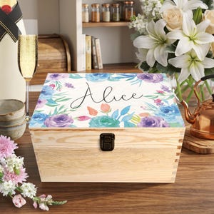 May include: A wooden box with a floral design and the name "Alice" on the lid. The lid features watercolor-style flowers in shades of blue, purple, and teal. The box has a metal clasp and is made of light-coloured wood.